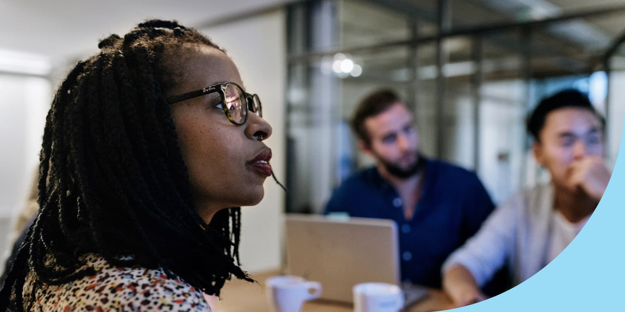 close-up shot of a Black woman sitting at a conference table looking to the side with two men sitting across the table from her visible in the background and an open laptop and mugs on the table
